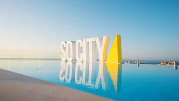 Une piscine dans laquelle le mot « SOCIÉTÉ » est inscrit en grandes lettres, entourée d’un ciel bleu clair et offrant une vue lointaine sur des bâtiments.