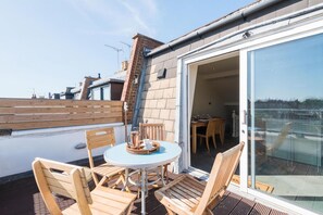 A rooftop terrace with a round table and wooden chairs, a glass door leading inside, and a view of neighboring buildings.