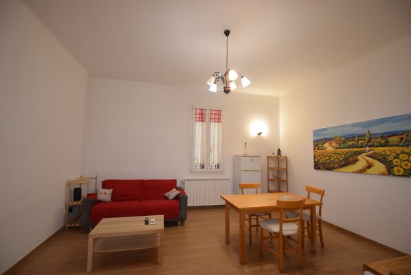 A living room with a red sofa, wooden dining table, and chairs. There is a window with curtains, a radiator, and a painting on the wall.