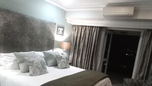 A hotel room with a large bed, a headboard, a nightstand, a lamp, a chair, and a view of the city at night. - 131 on Herbert Baker Boutique Hotel (Pretoria)