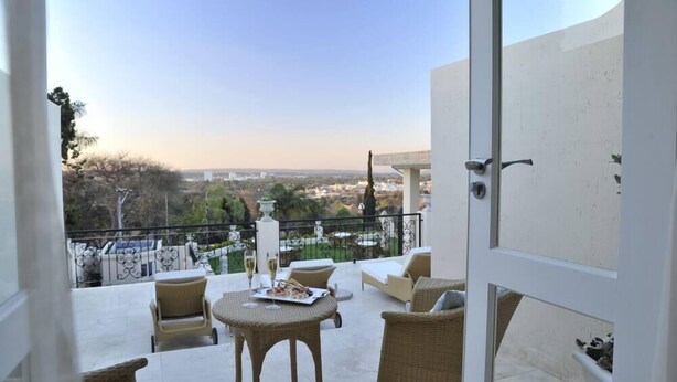 A balcony with wicker furniture, a table with a plate of food and a glass of wine, and a view of a cityscape. - 131 on Herbert Baker Boutique Hotel (Pretoria)