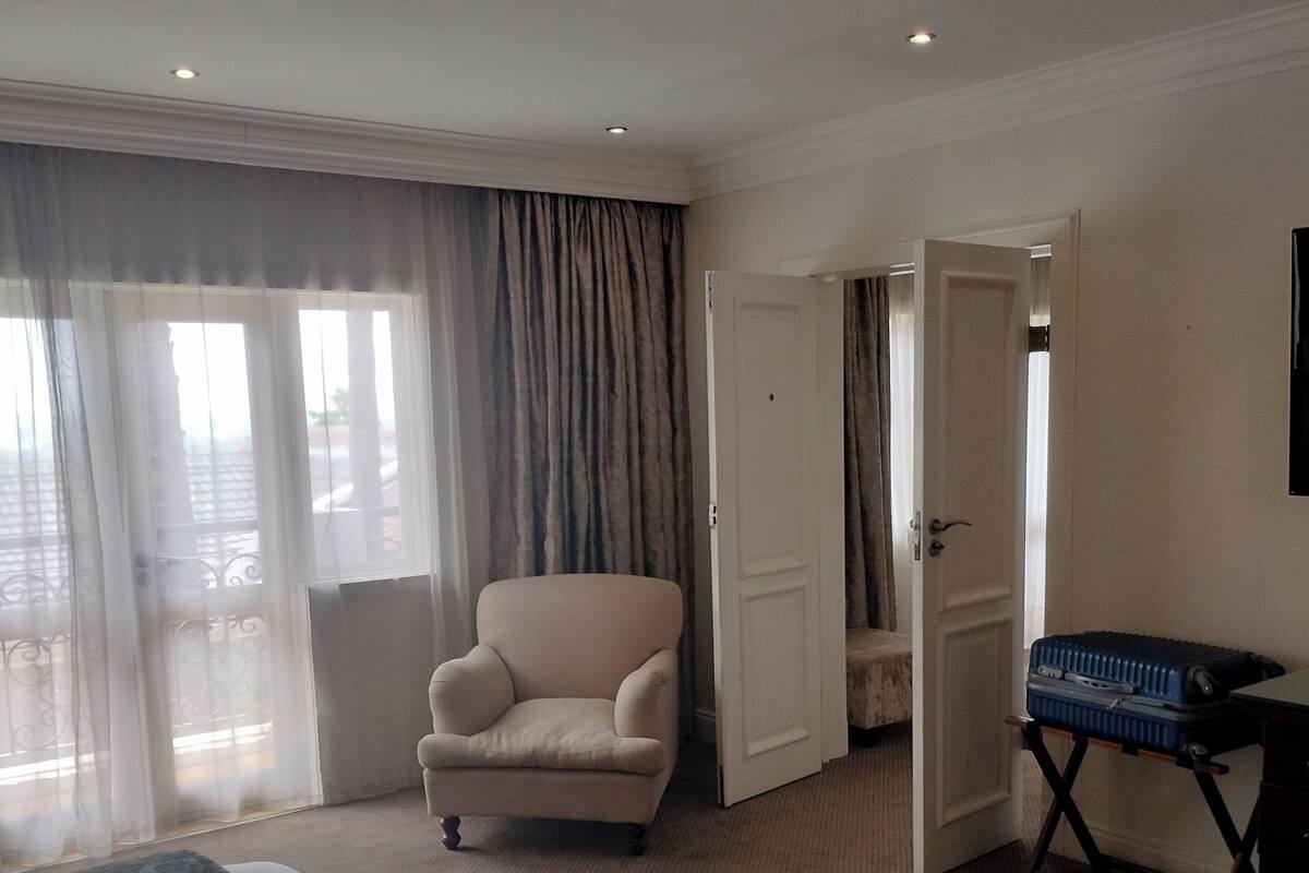 Ein Hotelzimmer mit einem großen Bett, einem Sessel, einem Schreibtisch mit einem Koffer und einer Tür zu einem anderen Raum.