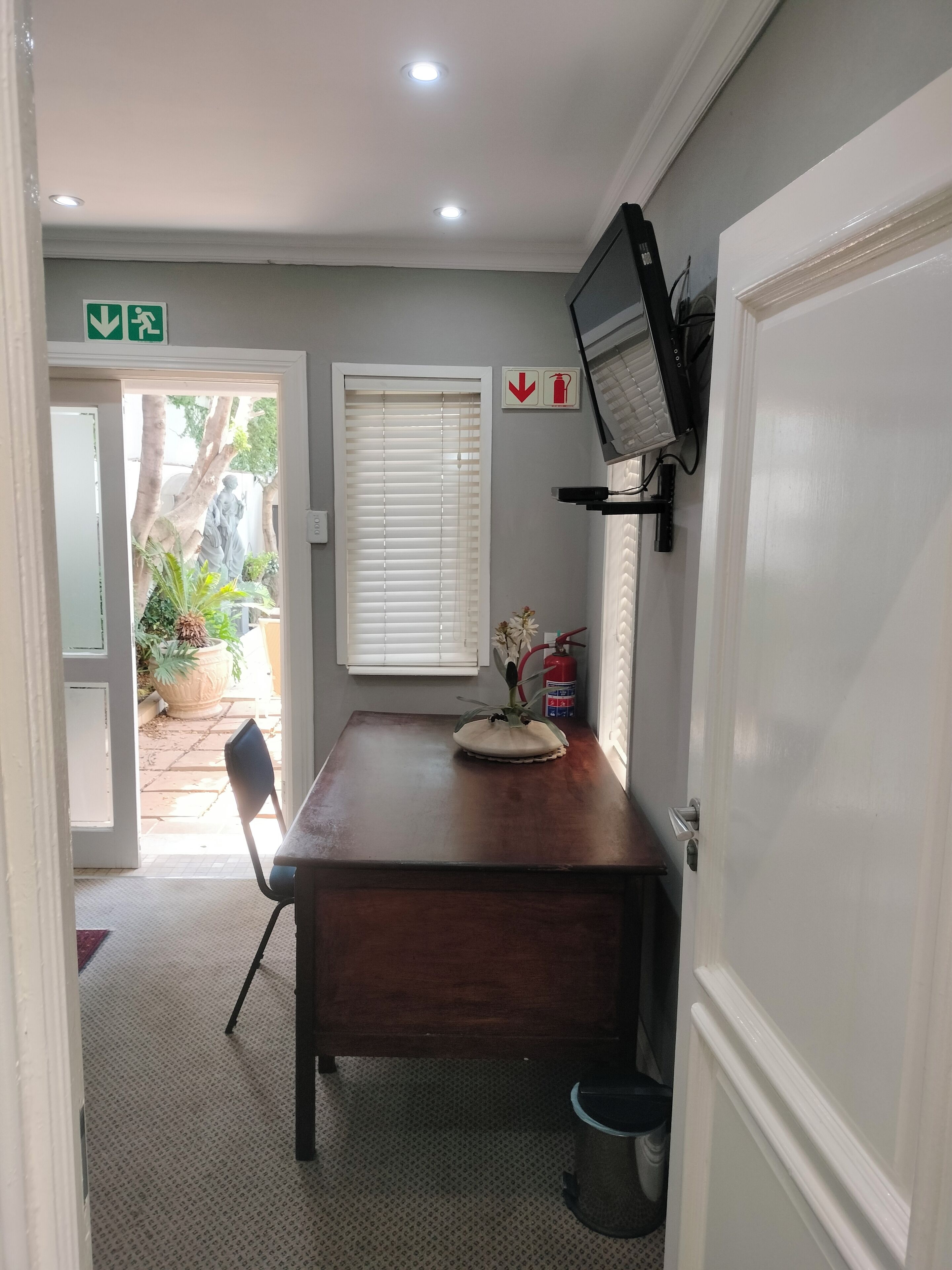 A room with a wooden table, a chair, a television mounted on the wall, a window with blinds, and a fire extinguisher.