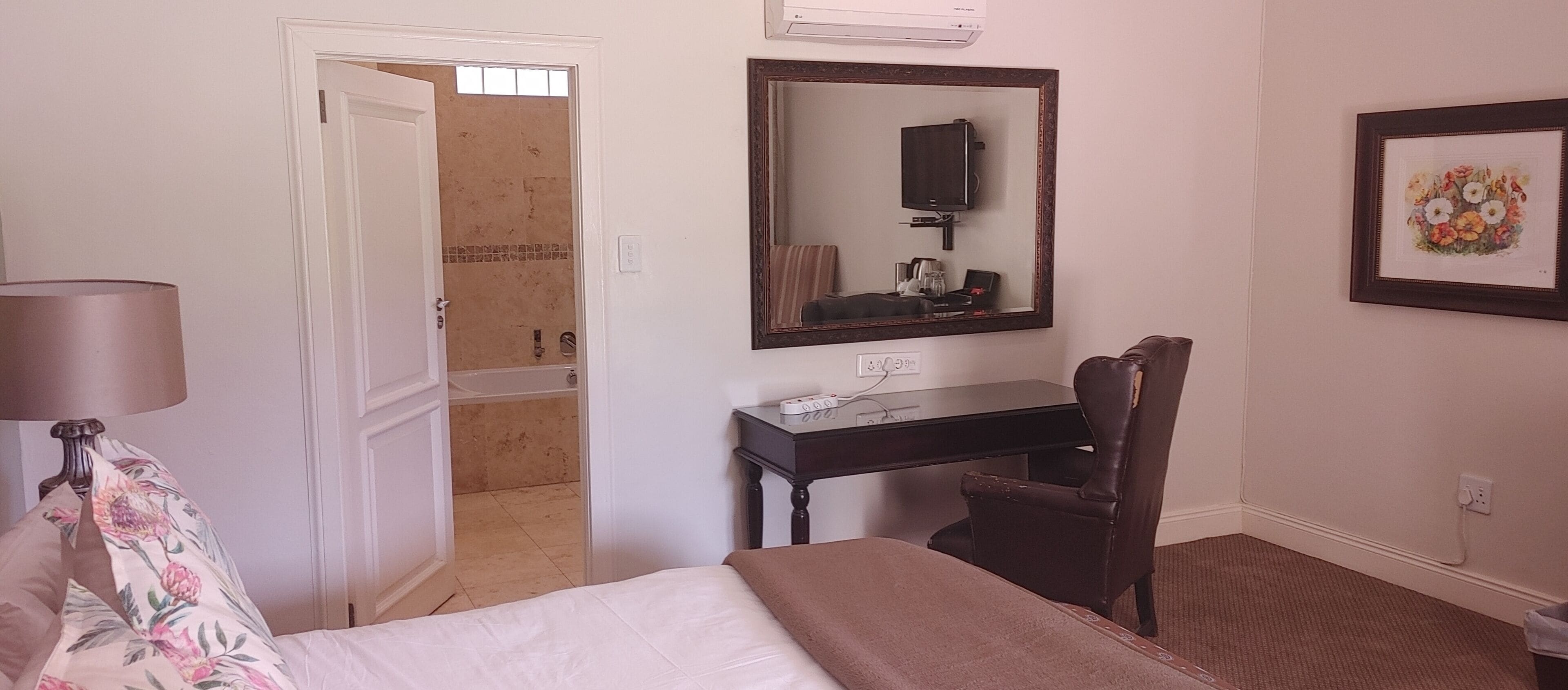 Quarto de hotel com uma cama, uma escrivaninha com cadeira, espelho, televisão e um quadro na parede.