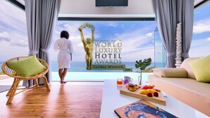 Ein Zimmer mit Meerblick, ein großes Fenster, ein Korbstuhl, ein Sofa und ein Frühstücksbrettchen.