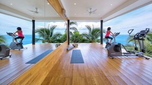 Ein geräumiger Fitnessraum mit Holzboden, Trainingsgeräten und großen Fenstern mit Blick auf den Ozean.