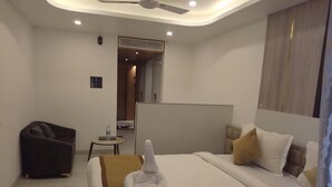 A hotel room with a bed, a chair, a small table with a bottle, and a ceiling fan.