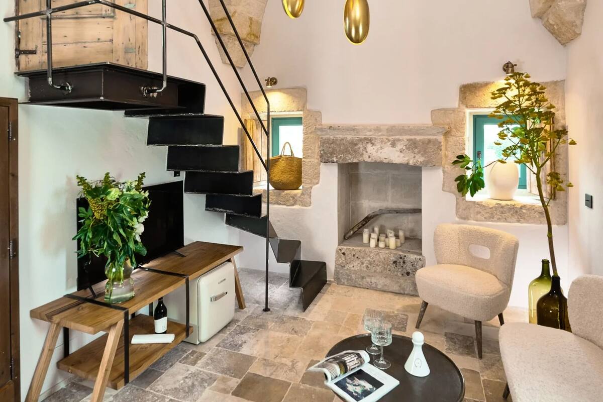 A modern living room with a stone fireplace, a wooden staircase, and hanging gold pendant lights.
