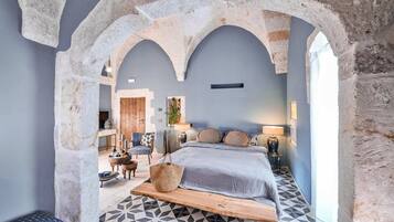 Quarto com arco de pedra, cama com colcha cinza, banco de madeira e chão estampado.