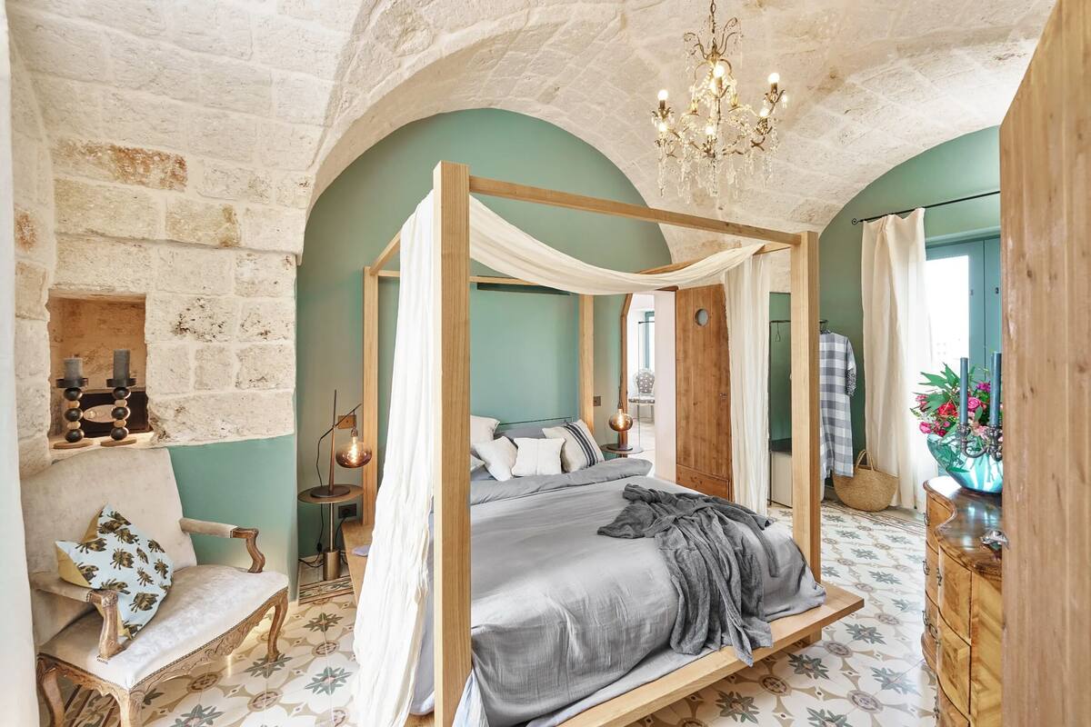 A four-poster bed with a canopy, a stone wall, a wooden chair, a small table with a lamp, and a mirror.