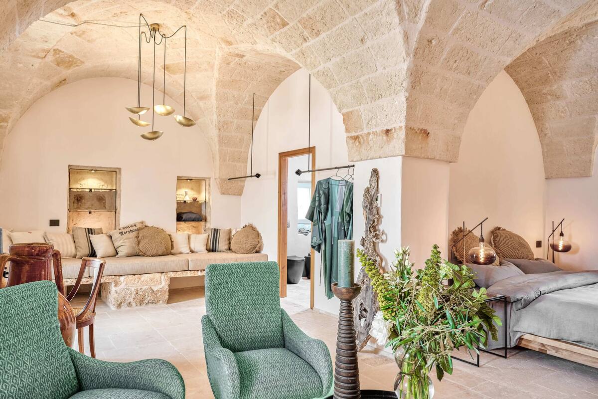 A cozy room with a stone arch ceiling, a green patterned armchair, a bed with a grey blanket, and a hanging plant.