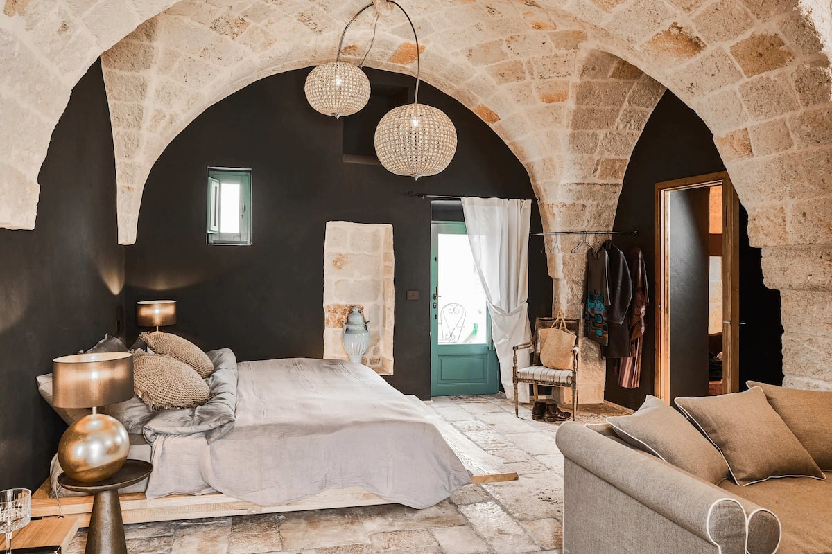A bedroom with a stone arch ceiling, a bed with a white cover, a grey sofa, and a hanging light fixture.