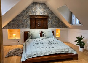 A bedroom with a sloped ceiling, a wooden headboard, a bed with a light blue bedspread, two bedside lamps, and a potted plant.