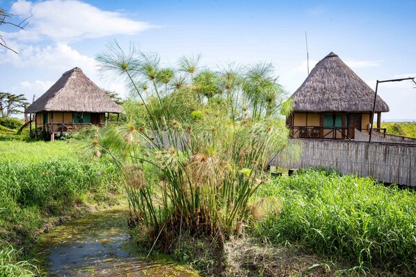 Two thatched-roof huts with wooden railings, a small stream, and lush greenery.