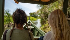 Dos personas en un bote, con un río y vegetación al fondo.