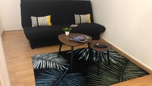 A small living area with a black sofa, two patterned cushions, a small round table with a magazine, a remote control, and a potted plant.