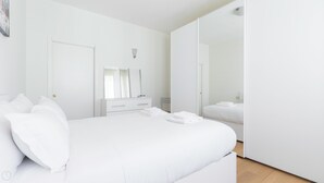 Une chambre à coucher moderne, décorée dans des tons de blanc, comprenant un grand lit, une commode, un miroir et une armoire.