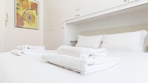Un lit bien fait, avec du linge de lit et des serviettes blanches, une tête de lit blanche et un tableau au mur.