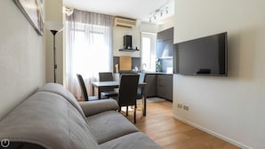 Een moderne woonkamer met een bank, een eettafel met stoelen, een keukenhoek met een aan de muur bevestigde televisie en een groot raam met gordijnen.