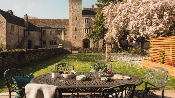 A round table with chairs and a tablecloth, set in a courtyard with a stone wall and a building in the background.