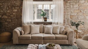 A cozy living room with a stone wall, a beige sofa, a window with white curtains, and a small side table with a plant.