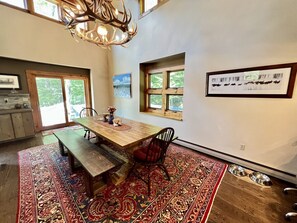 Un coin repas avec une table en bois, des bancs et un tapis rouge à motifs. Il y a des fenêtres donnant sur la verdure et un lustre au-dessus.
