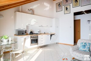 Una cucina moderna con mobili bianchi, un forno a microonde e un piano cottura. Si nota un tavolo da pranzo con sedie e un tavolo con ripiano in vetro. In primo piano si vede un divano con un cuscino floreale.