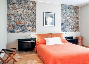 A bedroom with a stone wall, a bed with an orange bedspread, a nightstand, a piano, and a wicker chair.
