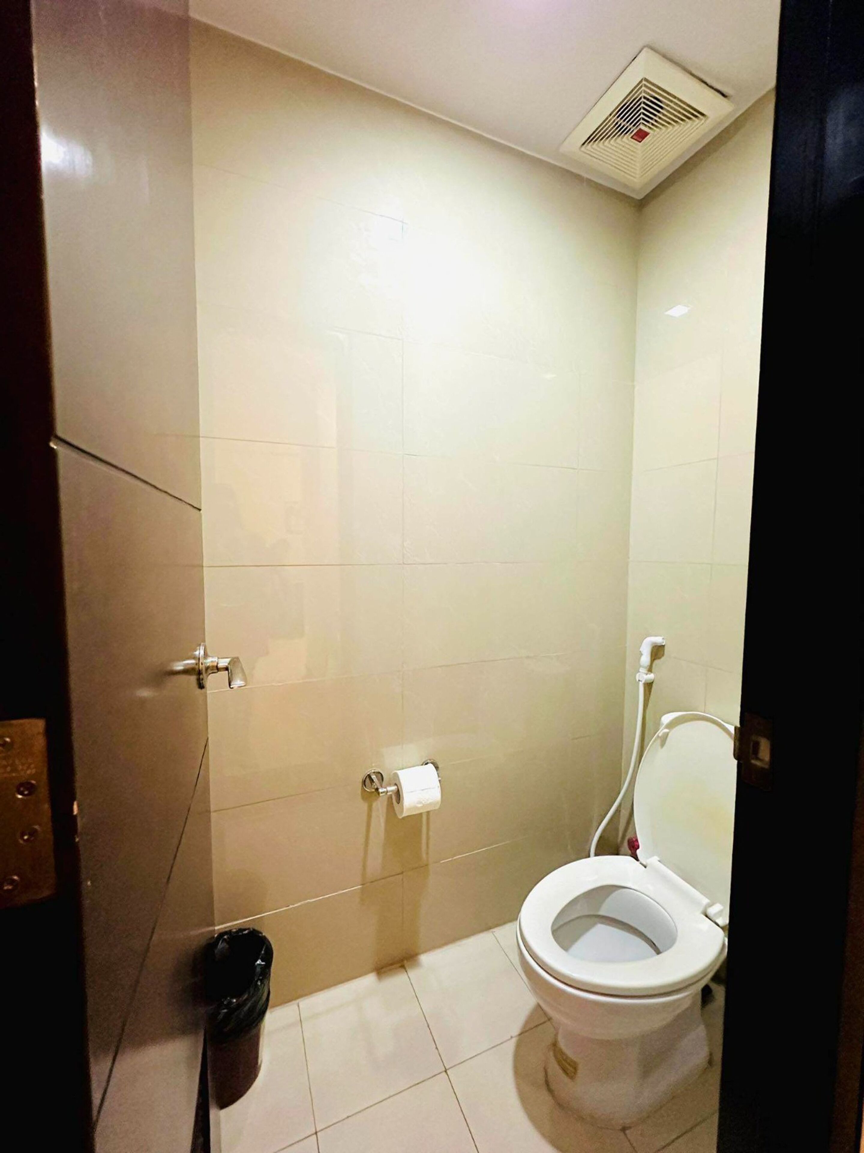 Un baño pequeño y bien iluminado con inodoro, un cesto de basura y una rejilla de ventilación en la pared.