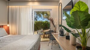 A modern bedroom with a large bed, a desk with chairs, a potted plant, and a view of the outdoors. - Amaryllis Hotel (Zakynthos)
