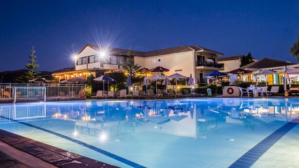 A swimming pool with lounge chairs and umbrellas, surrounded by buildings with outdoor seating and lights. - Amaryllis Hotel (Zakynthos)