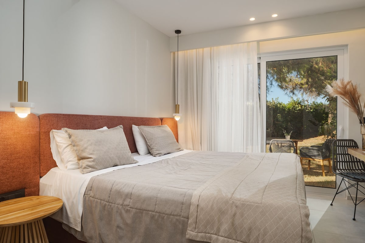 A modern bedroom with a large bed, a round wooden table, and a view of the outdoors through a sliding glass door.