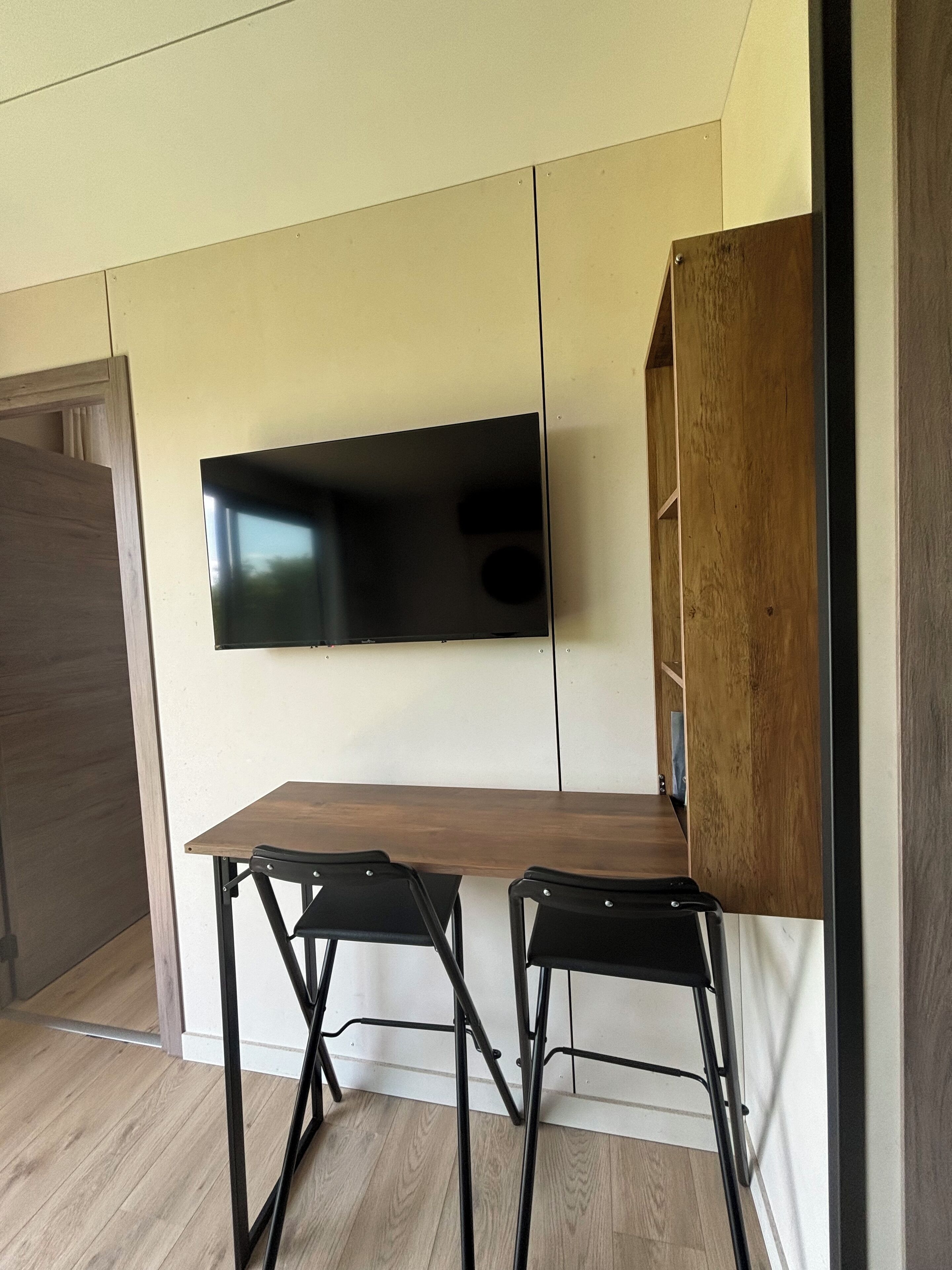 Een moderne kamer met een flatscreen televisie aan de muur, een houten bureau, twee zwarte stoelen en een houten kast.