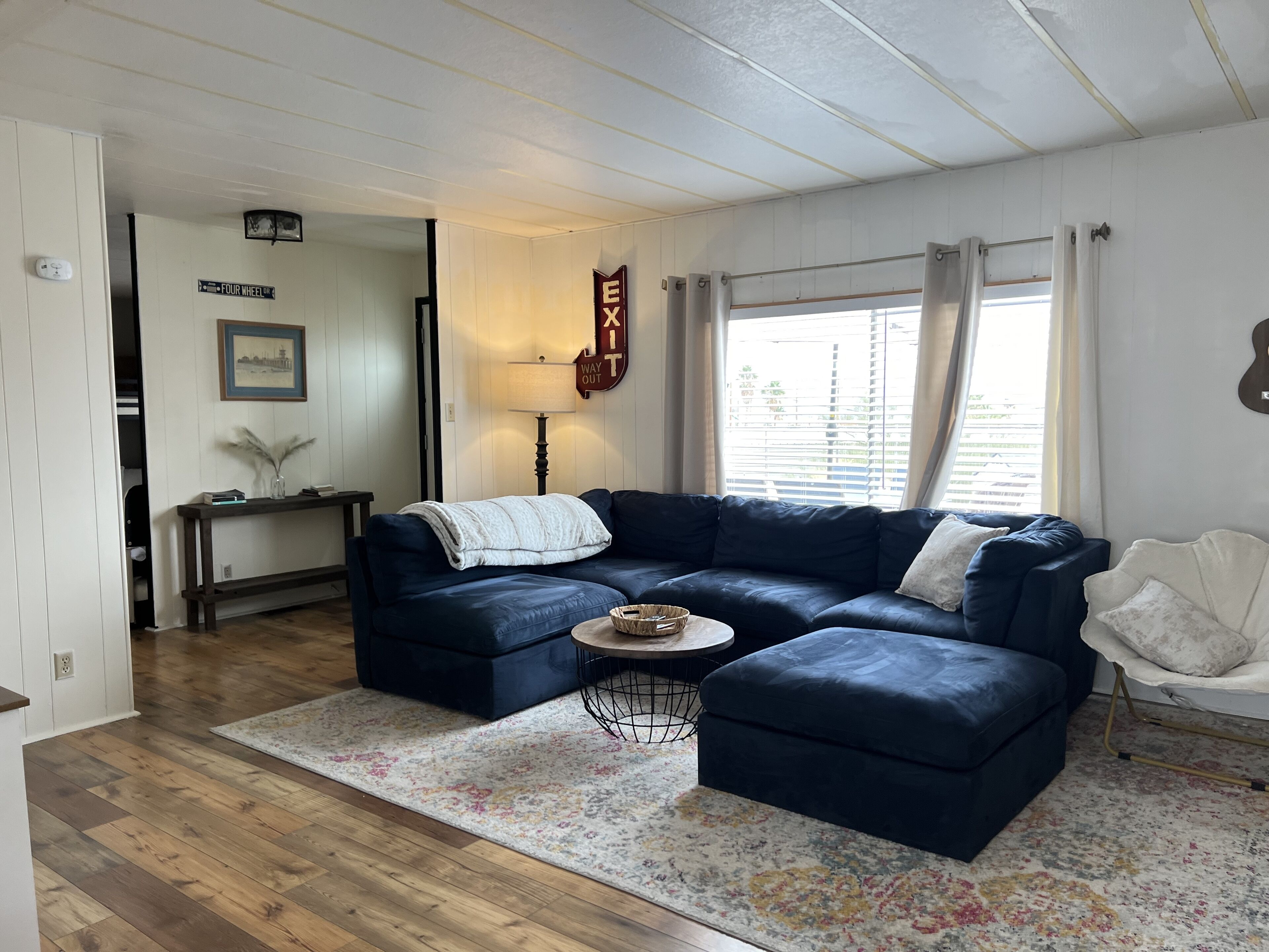 A living room with a dark blue sectional sofa, a round coffee table, and a patterned rug. There is a window with curtains, a lamp, and a wall-mounted sign.