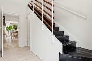 A modern staircase with wooden steps and white railings, leading to an open-plan living area with a dining table and chairs, and a view of greenery outside. - Rye Beach Chalet - Downstairs Only (Rye)
