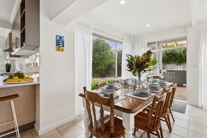 A dining area with a table set for six, surrounded by wooden chairs, a kitchen area with a counter, and a view of a garden through large windows. - Rye Beach Chalet and Loft (Rye)