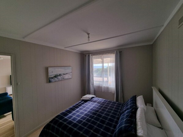 Un dormitorio con cama, ventana con cortinas, un cuadro en la pared y una puerta que da a otra habitación.