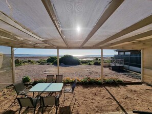 Een overdekt terras met een tafel en stoelen, met uitzicht op een zandrijk landschap met heuvels in de verte en een heldere lucht.