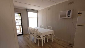 Un comedor con mesa y sillas blancas, piso de madera, ventana con persianas y un aire acondicionado instalado en la pared.