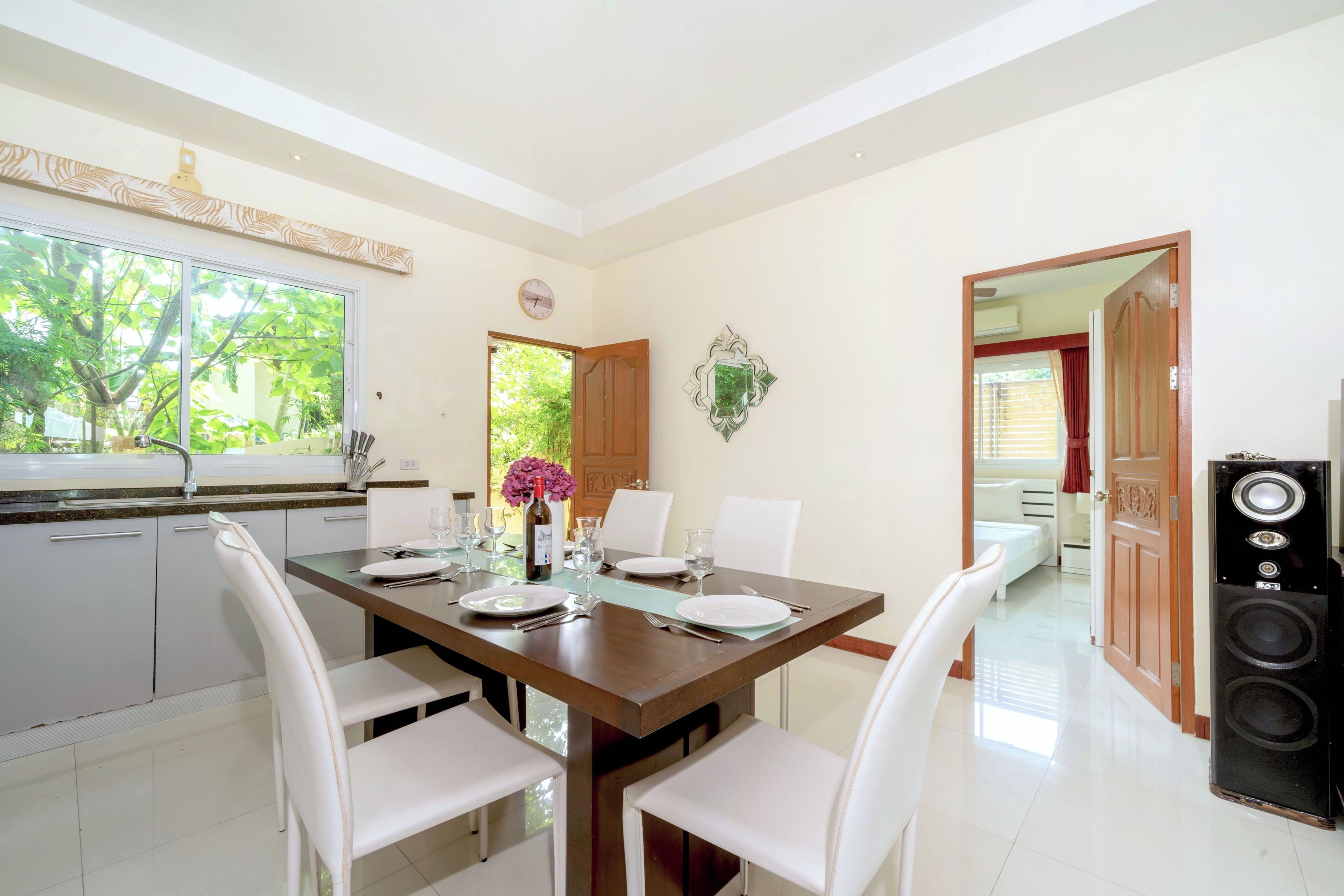 A dining area with a table set for four, white chairs, a clock on the wall, and a window with a view of trees.