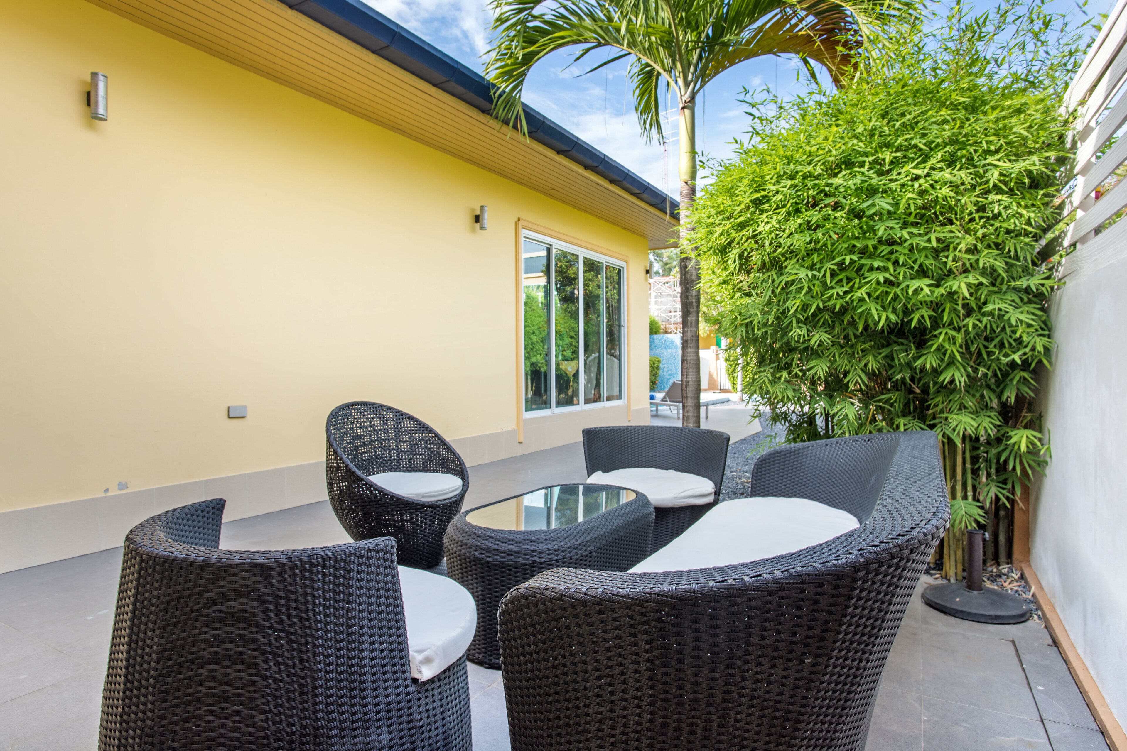 A patio with wicker furniture and a glass table, surrounded by a yellow wall and greenery.