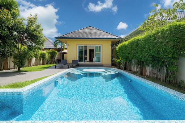 A swimming pool with a tiled edge, a covered patio area with chairs, and a house with a tiled roof and sliding glass doors.
