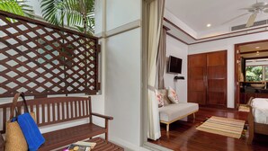 A room with a wooden bench, a small table with a book and a drink, a sofa with floral cushions, a TV mounted on the wall, and a wooden door leading to a bathroom. - Katamanda 3 Bed Tropical Villa B3 Near Kata Beach (Karon)