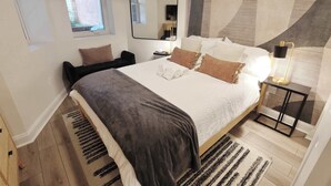 A bedroom with a bed, a bedside table, a lamp, a mirror, and a window.