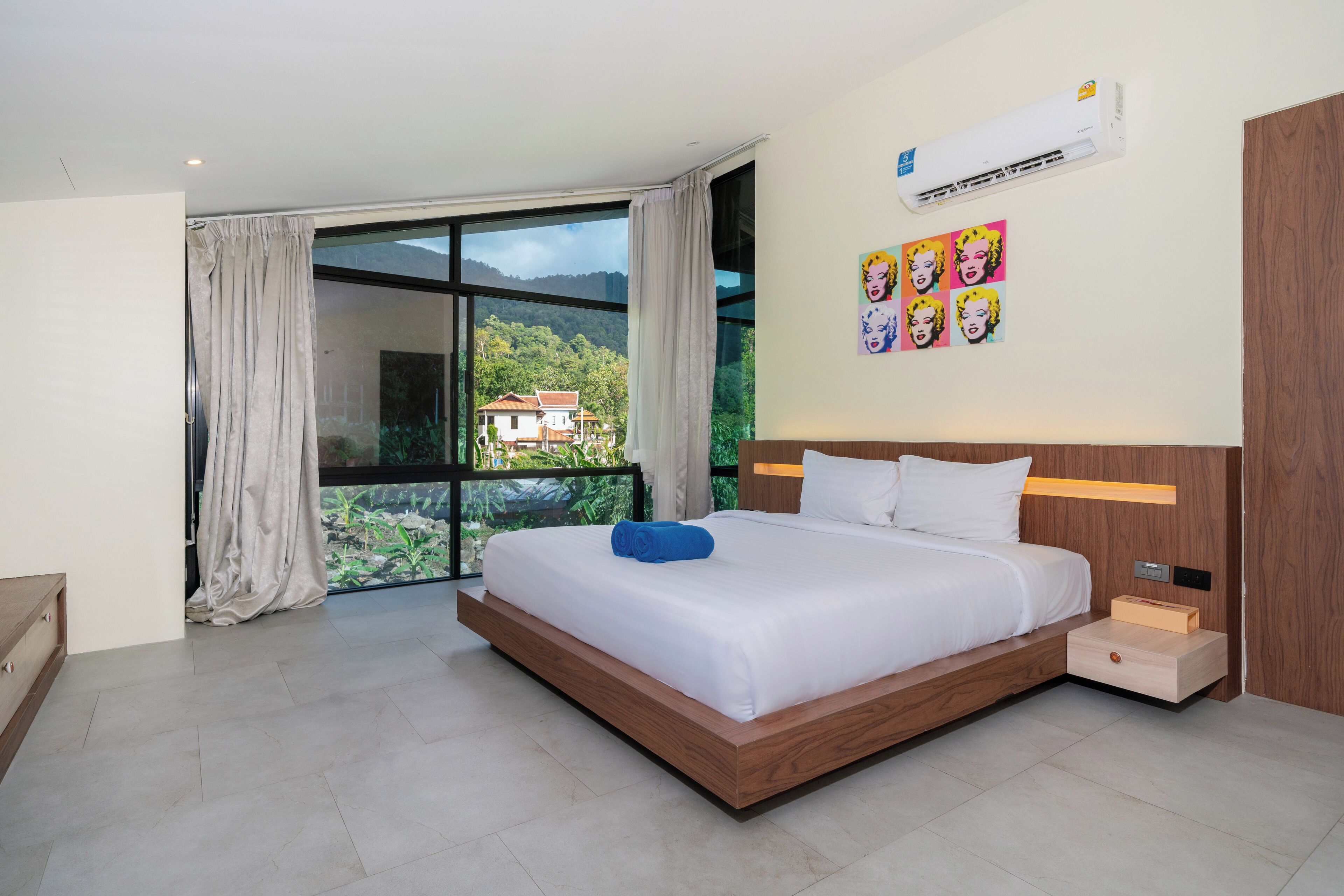 A modern hotel room with a large bed, a wooden headboard, an air conditioning unit, and a window with a view of greenery and a house.