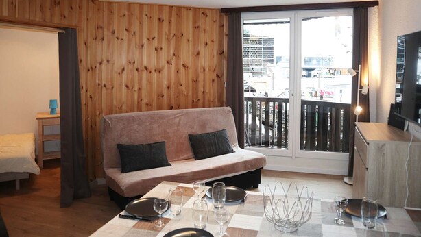 Ein modernes Wohnzimmer mit einer Holzvertäfelung, einer Couch, einem Esstisch gedeckt mit Tellern und Gläsern sowie einem Balkon mit Geländer.