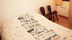 Ein Schlafzimmer mit einem Bett, das mit einer getupften Bettdecke bezogen ist, zwei Holzstühlen, einem Fenster mit gelben Vorhängen und einem Heizkörper.