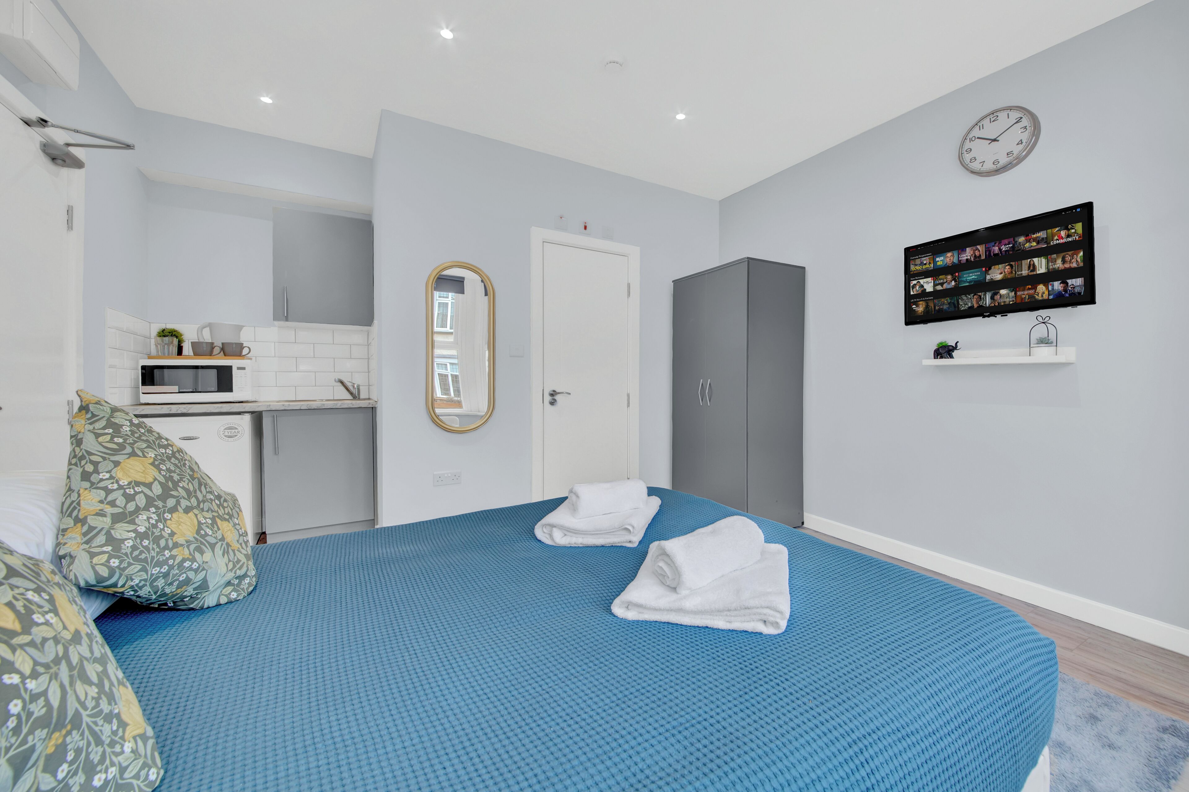 A modern bedroom with a blue bedspread, a white door, a grey wardrobe, a small shelf with a clock, and a TV on the wall.