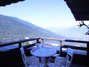 Um deck de madeira com uma mesa redonda e cadeiras brancas, com vista para uma paisagem montanhosa.