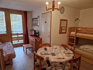 Um quarto acolhedor com uma cama de solteiro em madeira, uma escrivaninha com computador, uma mesa de jantar com cadeiras e uma janela com cortinas.